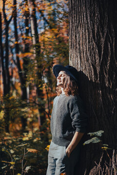 Vertical portrait of a dreamy young woman in a hat leaning against a tree trunk in autumn forest.