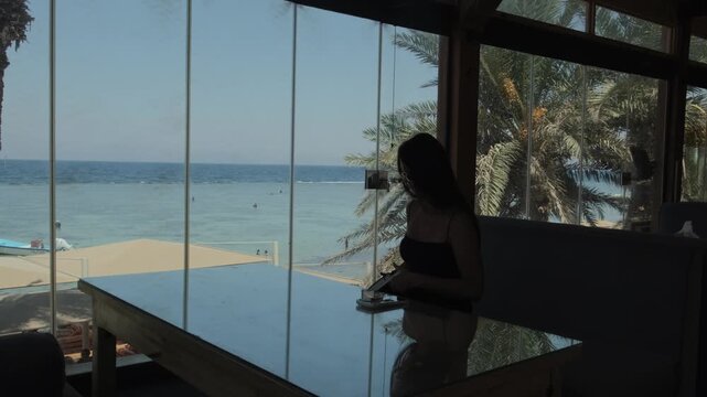 Woman arriving at seaside cafe, opening laptop and starting remote work with tropical sea and palm trees outside panoramic windows