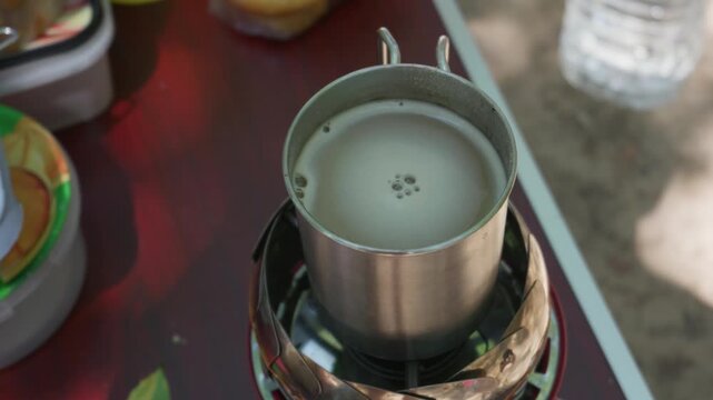 Metal mug filled with hot beverage boils on a portable gas stove during an outdoor camping trip. Concept of outdoor cooking, camping life, adventure travel, utility, and morning warmth.