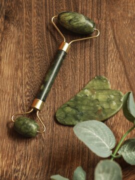 Jade facial roller and gua sha stone with eucalyptus leaves on wooden background