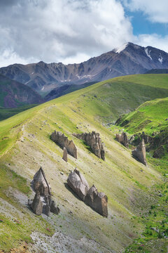 Sharp rock formations stand on a green mountain slope against a high peak under a cloudy sky. Vertical geological landscape