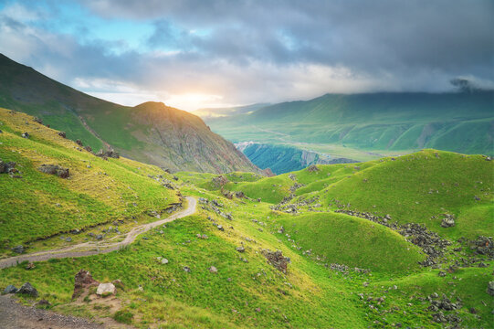 Winding dirt road cuts through green mountain slopes under a cloudy sunset sky with bright sunlight. Scenic alpine valley route