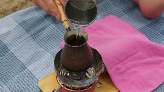 A hand pours water into an ornate copper cezve pot resting on a portable gas burner during an outdoor picnic. The small stove sits on a wooden board spread over a blue plaid blanket.
