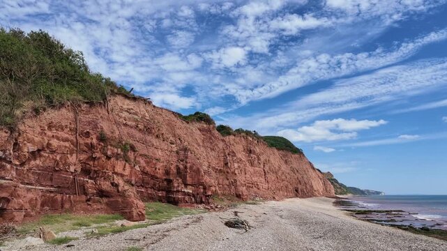 Sidmouth Devon England UK. 14.08.2026. Video. Expansive view of rugged, layered red sandstone cliffs plunging into the sea, with a sandy beach and a horizon stretching to distant headland
