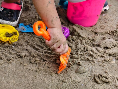 Close-up of a child's hand using an orange toy rake on sandy beach. Perfect for parenting, child development, family vacation, summer activities, and sensory play concepts.
