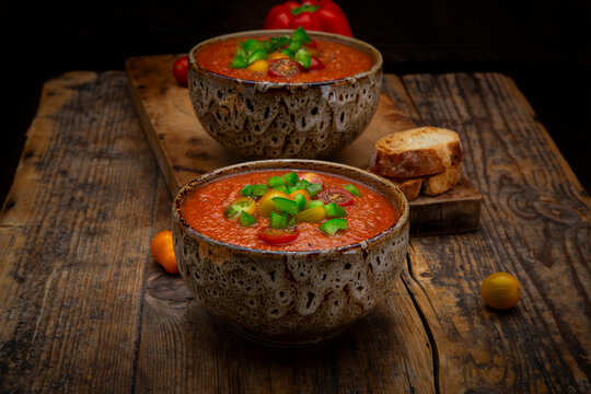Two bowls of cold Spanish gazpacho soup sit on a dark wooden table
