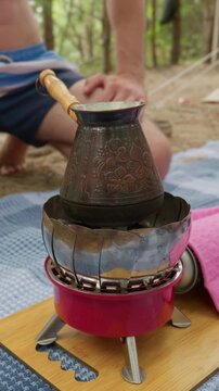 A decorated copper cezve heats on a portable gas stove at an outdoor campsite. Concept of outdoor lifestyle, rustic morning coffee preparation, relaxation, and wilderness camping.