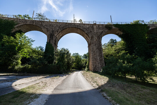 Historic stone arched railway bridge surrounded by green trees and ivy on a sunny day. A small rural road passes under the historic viaduct.