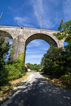 Historic stone arched railway bridge surrounded by green trees and ivy on a sunny day. A small rural road passes under the historic viaduct.