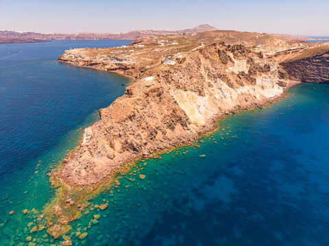 High altitude drone aerial view of rocky Cape Akrotiri surrounded by clear blue sea in Santorini
