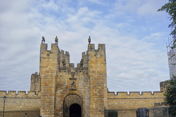 Majestic medieval stone castle fortress featuring ancient defense walls, battlements, and tall towers surrounded by green grass under a cloudy sky. Alnwick castle England, towers a very old castle © TomKorcak