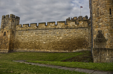 Majestic medieval stone castle fortress featuring ancient defense walls, battlements, and tall towers surrounded by green grass under a cloudy sky. Alnwick castle England, towers a very old castle © TomKorcak