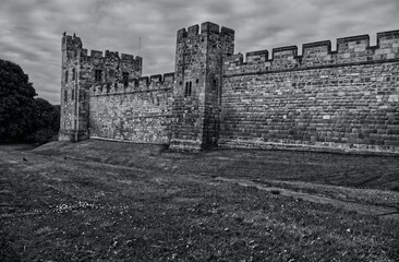Dramatic black and white landscape of a historic medieval stone castle fortress featuring defense walls, battlements, and towers under a cloudy sky. Alnwick castle England, towers a very old castle.  © TomKorcak