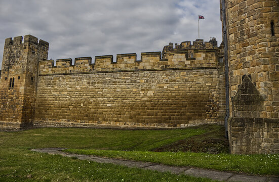 Majestic medieval stone castle fortress featuring ancient defense walls, battlements, and tall towers surrounded by green grass under a cloudy sky. Alnwick castle England, towers a very old castle