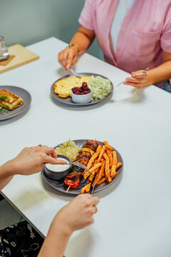 Two diners eat plated meals at a white restaurant table, including grilled meat with fries, sauce and cabbage, plus other dishes, with generous copy space