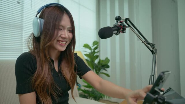 Asian female influencer smiles brightly and happily as she taps her smartphone to live singing on livestream. Concept of record cover song Freelance artist working from home ,social media creators.