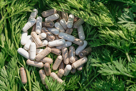 Close-up of white and clear herbal capsules with dietary supplements scattered, fresh green carrot tops. Healthy lifestyle, vegan vitamins, naturopathy, sports nutrition, and organic medical products