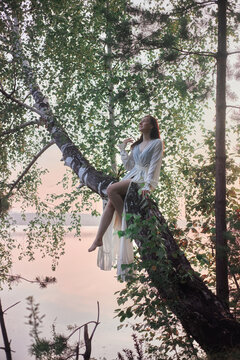 woman sits on a tree branch over water with a pink sky at sunrise. She wears a simple white robe and enjoys the nature around her, surrounded by leaves and branches