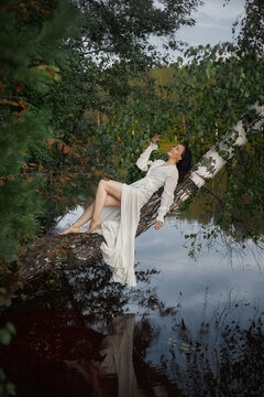 woman in a white dress sits on a tree branch over calm water. Green leaves surround her as she enjoys the natural setting. sun is low, creating soft light