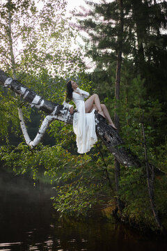 woman with long dark hair sits on a tree branch above still water. She wears a white dress and looks thoughtful. background has green trees and a quiet atmosphere