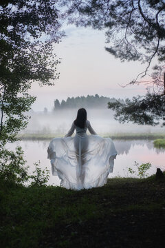 woman stands by a lake at dawn wearing a white dress. Fog covers the water and trees. sun starts to rise, casting soft light over the scene. Nature surrounds her