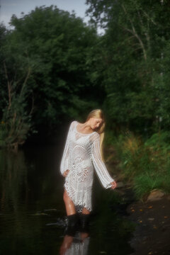 A woman stands in shallow water surrounded by trees. She wears a long light-colored dress and has a cheerful expression. The day appears cloudy and the atmosphere is natural