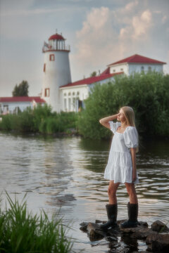 A girl in a white dress poses on rocks in the water. A lighthouse and buildings are visible in the background. There are green plants around and the sky is clear
