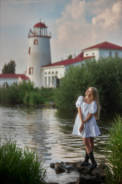 A young woman wears a white dress and black boots while standing on rocks beside a river. The lighthouse and buildings are visible in the background under a cloudy sky