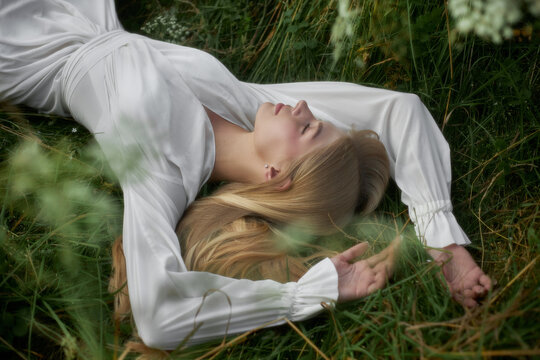 A woman lies on the grass in a field under a clear sky. She has long hair and is wearing a white dress. Bright green plants surround her while she enjoys the moment