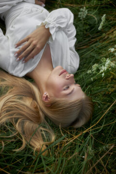 A woman with long hair rests on green grass in a field. She wears a white top and gazes upwards. It is daytime and the sun is shining bright