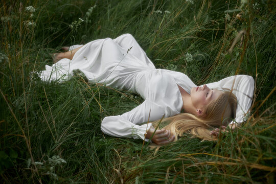 A woman lies on her back in a field of grass wearing a long white dress. The sun shines down, and she has her eyes closed, enjoying the moment outdoors in nature