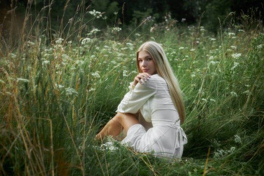 A young woman poses in a grass field wearing a white dress. The sun shines as she sits among tall green grass and small flowers. The scene shows nature and a clear day