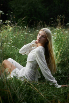 A young woman is seated in tall grass among white wildflowers. She is wearing a light outfit and has long hair. The scene is bright and captures a natural moment in nature
