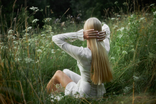 A woman sits among tall grass and wildflowers with her back to the viewer. She is arranging her long hair while enjoying the nature around her in a sunny environment