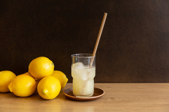 Citronnade bien fra&icirc;che avec des gla&ccedil;ons, servie dans un grand verre transparent avec une paille brune, table en bois, fond brun fonc&eacute;, citrons jaunes entiers empil&eacute;s &agrave; gauche du verre 