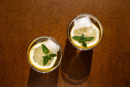 Deux verres transparents remplis avec de l'eau p&eacute;tillante, des rondelles de citron jaune, des feuilles de menthe et des gla&ccedil;ons, fond brun, vue du dessus, boisson rafraichissante