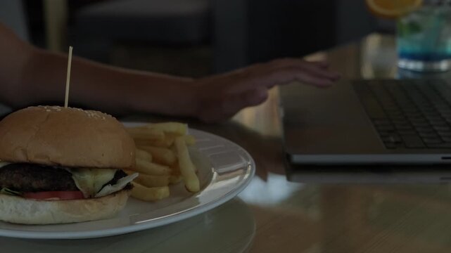 Close up of burger and french fries beside laptop while woman works at seaside cafe showing unhealthy work habits sedentary lifestyle eating during work and lack of breaks