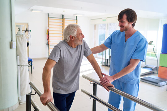 Senior Man Using Parallel Bars With Physiotherapist Assistance In Rehab Center For Mobility And Strength Training Indoors