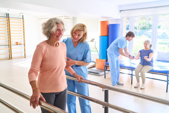 Senior Woman Walking With Parallel Bars Assisted By Physiotherapist In Rehab Center For Rehabilitation And Recovery After Injury Indoors