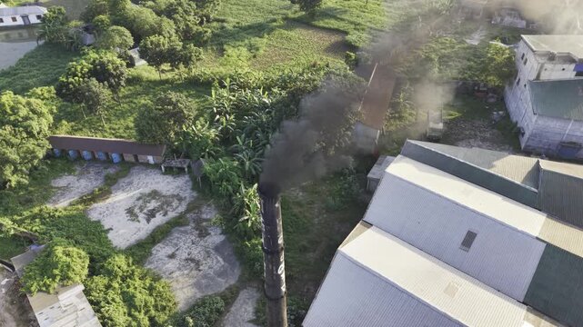 Drone footage showing an industrial smokestack and smoke plume surrounded by green fields, trees and countryside homes, suitable for environmental awareness, industry, agriculture, and sustainability 