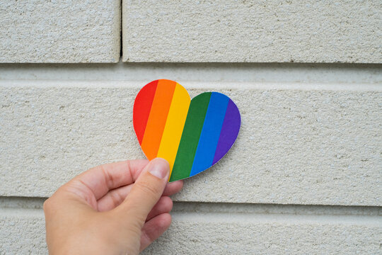 Hand holding rainbow heart symbol on light wall background for LGBTQ pride and equality concept	
