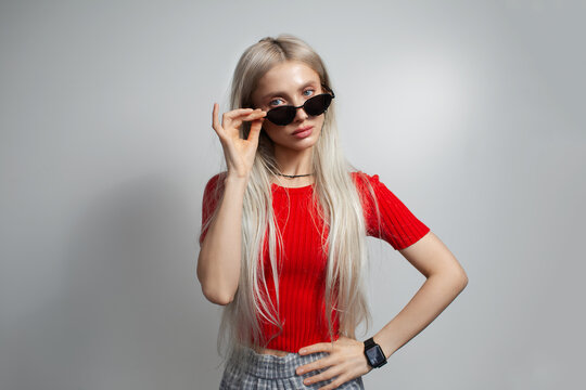 Young pretty blonde girl in red shirt wearing sunglasses isolated on grey background.