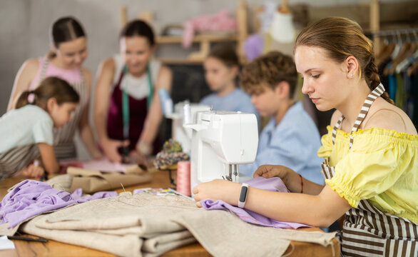 Smiling teen girl focusing on sewing on machine during hands-on lesson while classmates preparing patterns, fabric and fitting garments with female teacher