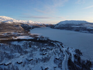 Stunning high-angle aerial view of a frozen lake surrounded by snow-covered mountains and remote wooden cabins in a winter landscape at dawn. © nikjuzaili
