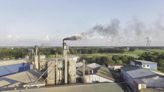 Aerial view of Khajanagar Rice Mill in Kushtia, Bangladesh, showing industrial processing structures and visible smoke beside green farmland, power lines and rural landscape from above.