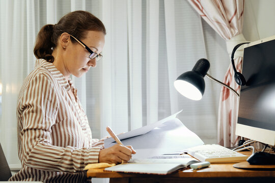 Focused middle-aged woman wearing glasses sits her home desk reading and writing paper documents under light of desk lamp, next to computer monitor, in evening.