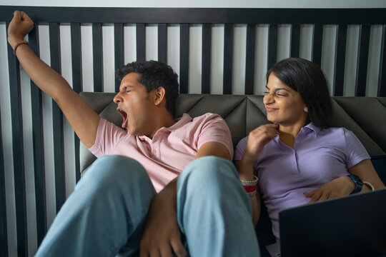 A young newlywed couple in bedroom with the husband yawning and wife using laptop.