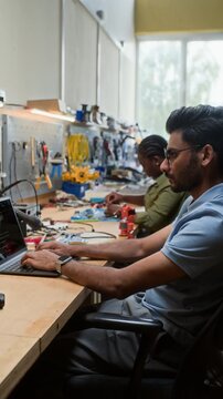Vertical video of Indian young adult man coding on laptop while African American young adult woman assembling hardware. Footage is perfect for STEM education and robotics engineering ads