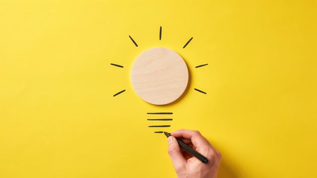 A hand using a black marker to draw a light bulb outline around a circular wooden disc placed on a vibrant yellow paper background surface