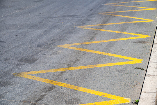 Bright yellow zig-zag road markings painted on a textured grey asphalt street next to a concrete curb, indicating a restricted parking or no-stopping zone.